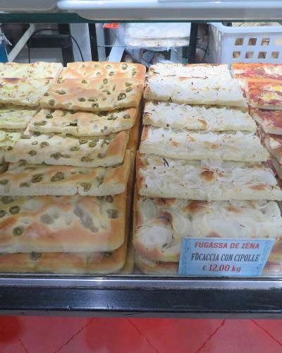 A display case in a bakery or food shop showing stacks of various rectangular flatbreads, including one labeled "FUGÀSSA DE ZÉNA" (Focaccia with onions) priced at €12.00/kg.