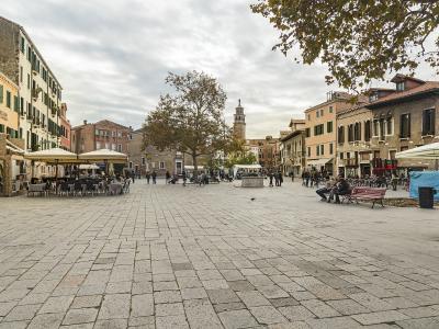 A wide, paved public square in Venice featuring outdoor cafe seating, a central stone well, a bell tower in the background, and surrounding multi-story buildings with shuttered windows.