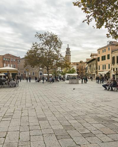 A wide, paved public square in Venice featuring outdoor cafe seating, a central stone well, a bell tower in the background, and surrounding multi-story buildings with shuttered windows.