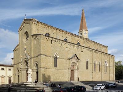 A large Romanesque-Gothic style stone cathedral with a tall brick spire, situated on a wide set of steps in a town square with parked cars and pedestrians.