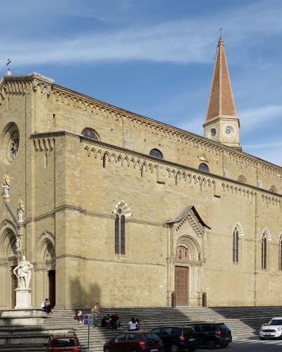 A large Romanesque-Gothic style stone cathedral with a tall brick spire, situated on a wide set of steps in a town square with parked cars and pedestrians.