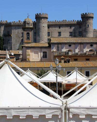 A large medieval-style stone castle with prominent round towers and crenellated battlements, situated on a hill above a town square filled with white event tents under a clear blue sky.