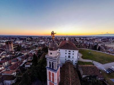 An aerial view of a historic Italian hilltop complex featuring a prominent bell tower topped with a golden statue of an angel, overlooking a dense urban landscape at sunset.
