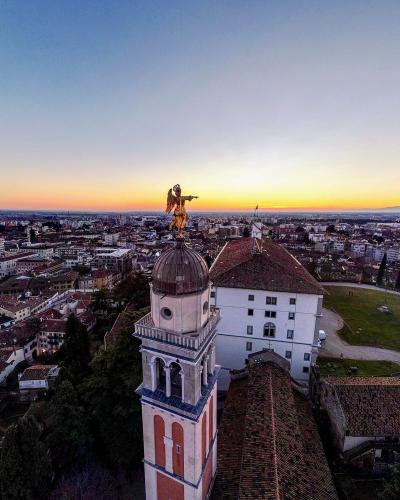 An aerial view of a historic Italian hilltop complex featuring a prominent bell tower topped with a golden statue of an angel, overlooking a dense urban landscape at sunset.