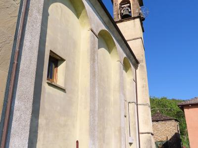 The image shows the exterior side and bell tower of a church building with a pale yellow stucco facade, arched blind arcades, and a small bell gable, set against a blue sky with green grass in the foreground.