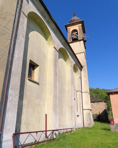 The image shows the exterior side and bell tower of a church building with a pale yellow stucco facade, arched blind arcades, and a small bell gable, set against a blue sky with green grass in the foreground.