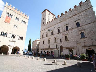 The image shows a large, paved public square flanked by two prominent medieval stone buildings with crenellated roofs; the building on the left features a banner for "MUSEO CIVICO DI TUDER" and the one on the right has a "FARMACIA" sign, with a st