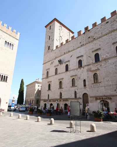 The image shows a large, paved public square flanked by two prominent medieval stone buildings with crenellated roofs; the building on the left features a banner for "MUSEO CIVICO DI TUDER" and the one on the right has a "FARMACIA" sign, with a st
