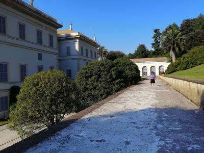 A sunlit view of a large, pale-colored neoclassical villa with shuttered windows and a terraced garden path leading toward a colonnaded structure, with a visitor walking along the path under a clear blue sky.