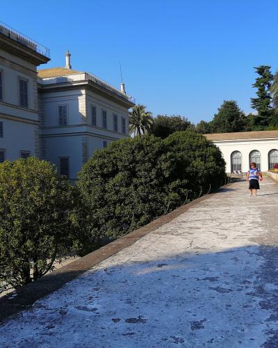 A sunlit view of a large, pale-colored neoclassical villa with shuttered windows and a terraced garden path leading toward a colonnaded structure, with a visitor walking along the path under a clear blue sky.