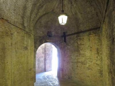 A dimly lit, arched brick tunnel with a hanging lantern and a bright arched exit at the far end, showing worn brick walls and a ribbed stone floor.