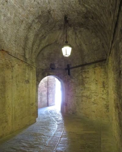 A dimly lit, arched brick tunnel with a hanging lantern and a bright arched exit at the far end, showing worn brick walls and a ribbed stone floor.