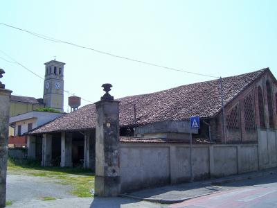 A street-level view of a long, single-story brick building with a tiled roof and arched windows, fronted by a concrete wall and stone pillars, with a church bell tower visible in the background.