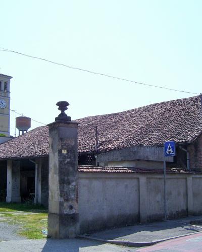 A street-level view of a long, single-story brick building with a tiled roof and arched windows, fronted by a concrete wall and stone pillars, with a church bell tower visible in the background.