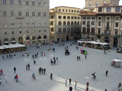 A high-angle view of a large, paved public square surrounded by multi-story buildings with Renaissance architecture, featuring outdoor cafes, pedestrians, and a horse-drawn carriage.