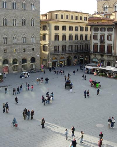 A high-angle view of a large, paved public square surrounded by multi-story buildings with Renaissance architecture, featuring outdoor cafes, pedestrians, and a horse-drawn carriage.