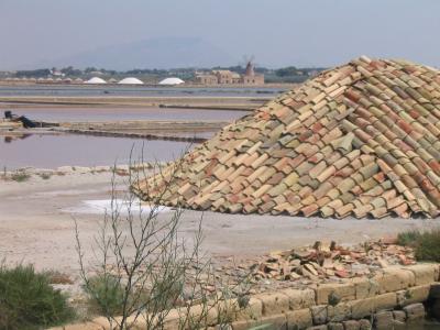 A large conical pile of terracotta roof tiles in the foreground of a salt flat landscape, with pinkish water channels, distant white salt mounds, and a traditional windmill building visible in the background.