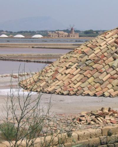 A large conical pile of terracotta roof tiles in the foreground of a salt flat landscape, with pinkish water channels, distant white salt mounds, and a traditional windmill building visible in the background.
