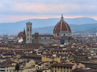 A twilight view of the Florence Cathedral (Duomo) and its bell tower illuminated against a dusky sky, seen from an elevated vantage point over the city's rooftops with hills in the background.