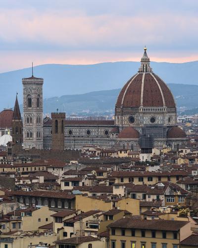 A twilight view of the Florence Cathedral (Duomo) and its bell tower illuminated against a dusky sky, seen from an elevated vantage point over the city's rooftops with hills in the background.