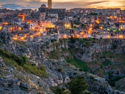 A twilight view of the Sassi di Matera district, featuring the illuminated Cathedral of Matera and its bell tower on a rocky plateau above a deep ravine.