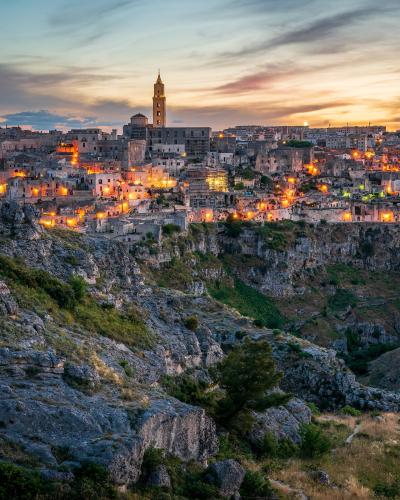 A twilight view of the Sassi di Matera district, featuring the illuminated Cathedral of Matera and its bell tower on a rocky plateau above a deep ravine.