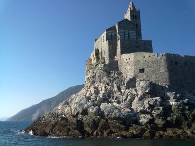 The image shows the Church of San Pietro perched on a rocky promontory overlooking the sea, with a distinctive striped facade and bell tower, located in Portovenere, Italy.