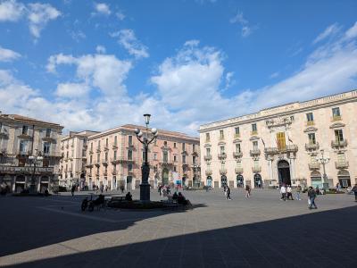A wide-angle daytime view of a large, paved public square in Catania, Italy, featuring the University of Catania building on the right with "UNIVERSITAS STUDII CATANIENSIS" inscribed above its entrance, flanked by other historic buildings and a ce