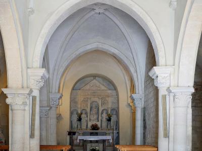 The interior of a historic stone church featuring a nave with wooden pews, pointed arches, and an altar area with a sculpted reredos depicting religious figures.