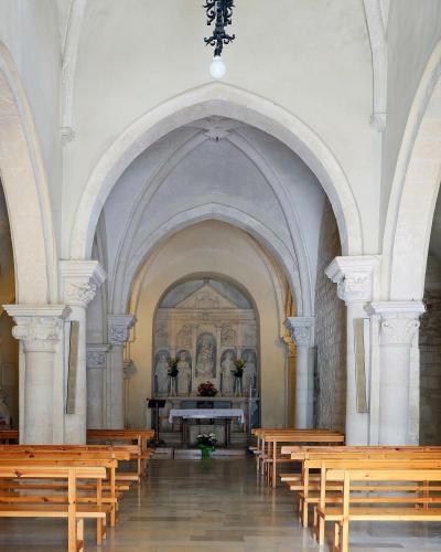 The interior of a historic stone church featuring a nave with wooden pews, pointed arches, and an altar area with a sculpted reredos depicting religious figures.