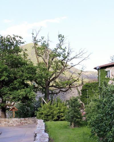A paved path leading past stone walls and lush greenery towards a stone building with a tiled roof, set against a backdrop of a forested hill.