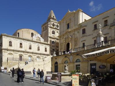 A sunlit view of a grand Baroque church complex with a prominent bell tower and a wide staircase leading up to it, situated in a public square with pedestrians and a restaurant awning in the foreground.