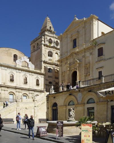 A sunlit view of a grand Baroque church complex with a prominent bell tower and a wide staircase leading up to it, situated in a public square with pedestrians and a restaurant awning in the foreground.