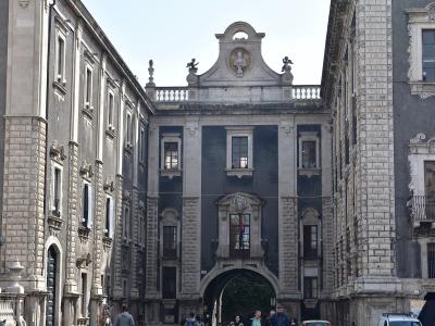 A Baroque stone archway connecting two buildings, featuring a central bust in a pediment, statues on the balustrade, and an inscription reading "D O SCIENTIA ET VIRTUTE DECUS" above the arch, with pedestrians and vehicles at street level.