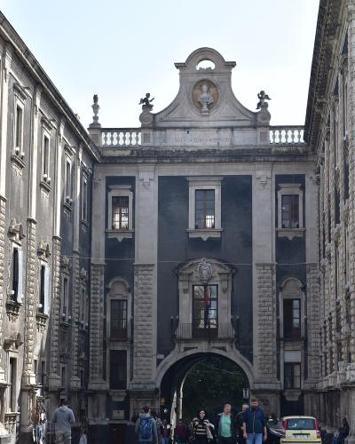 A Baroque stone archway connecting two buildings, featuring a central bust in a pediment, statues on the balustrade, and an inscription reading "D O SCIENTIA ET VIRTUTE DECUS" above the arch, with pedestrians and vehicles at street level.
