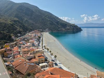 A high-angle view of a long, sandy beach curving along a coastal town with terracotta-roofed buildings, palm trees lining the shore, and a steep green mountain rising behind the town, under a blue sky with scattered clouds.