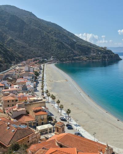 A high-angle view of a long, sandy beach curving along a coastal town with terracotta-roofed buildings, palm trees lining the shore, and a steep green mountain rising behind the town, under a blue sky with scattered clouds.
