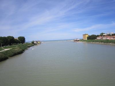 A wide river mouth flowing into the sea under a blue sky, with a paved path and trees on the left bank and buildings on the right bank.