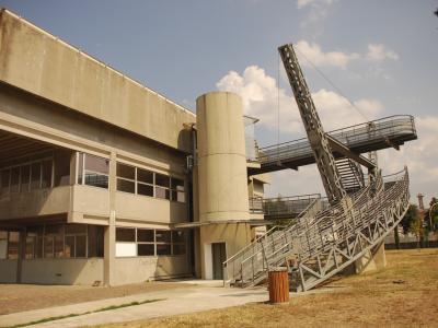 A concrete building with a large, distinctive metal staircase structure leading to an elevated platform, set in a grassy outdoor area under a blue sky.