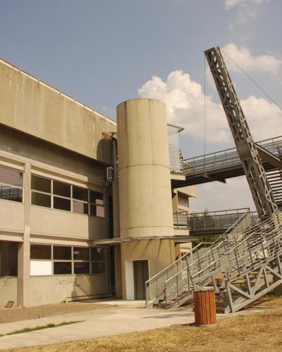 A concrete building with a large, distinctive metal staircase structure leading to an elevated platform, set in a grassy outdoor area under a blue sky.