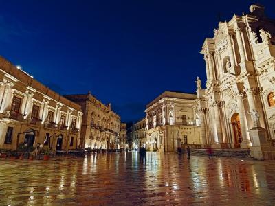 A wide, wet-paved European square at twilight, flanked by illuminated Baroque-style buildings and dominated on the right by a large, ornate cathedral facade with statues and columns.
