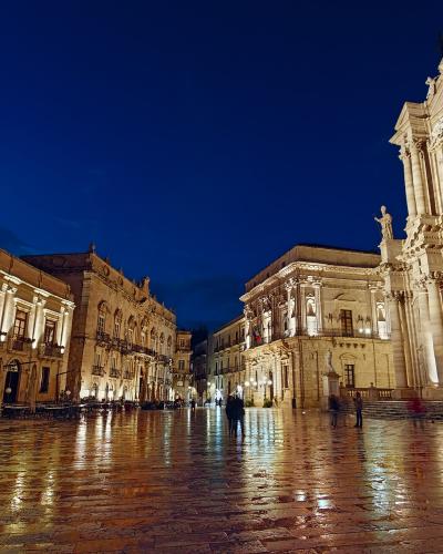 A wide, wet-paved European square at twilight, flanked by illuminated Baroque-style buildings and dominated on the right by a large, ornate cathedral facade with statues and columns.