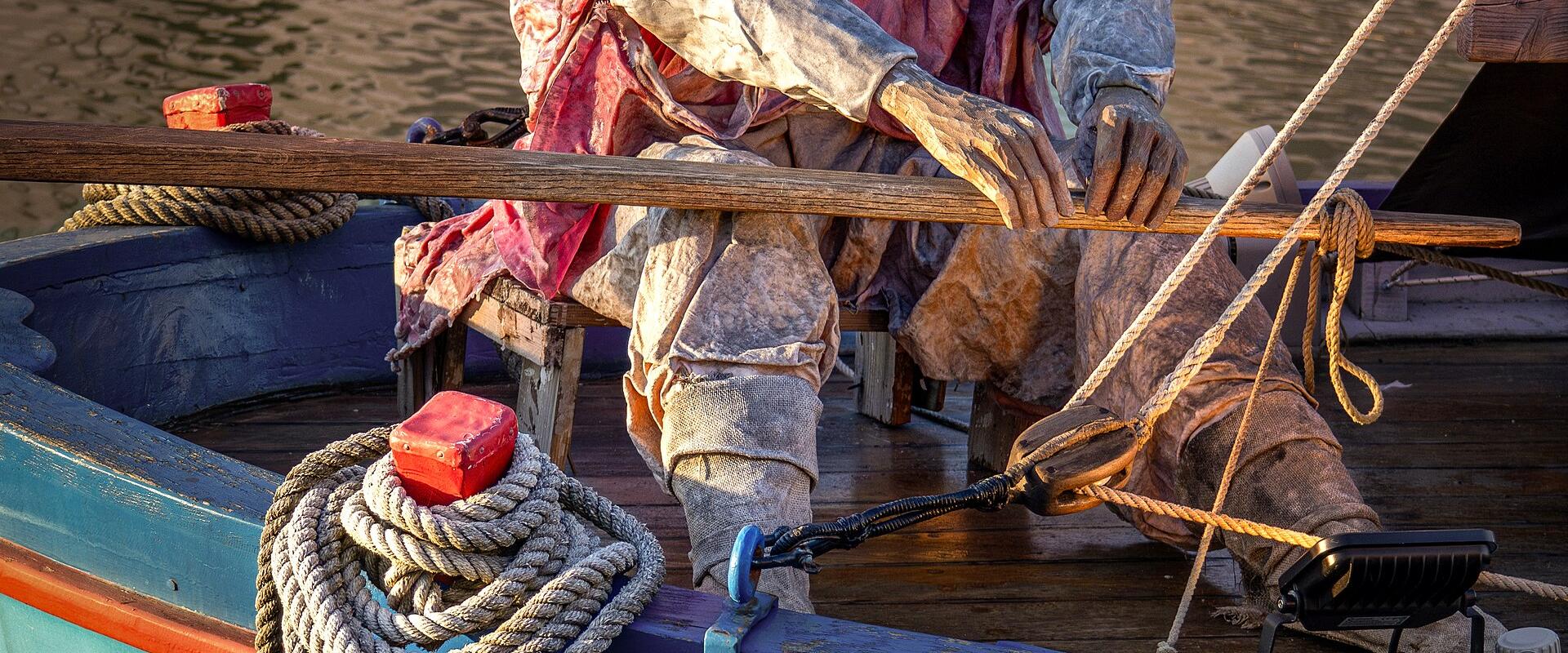 A life-sized statue of a fisherman seated in a small, painted wooden boat named "Lancia," moored in calm water with ropes and rigging visible.