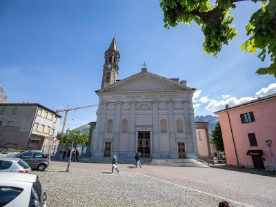 A white neoclassical church with a prominent bell tower and the Latin inscription "DIVO NICOLAO LEUCI PATRONO" on its facade, situated in a paved town square with mountains in the background.