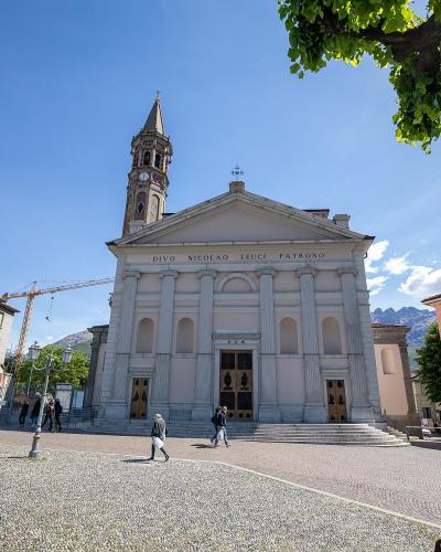 A white neoclassical church with a prominent bell tower and the Latin inscription "DIVO NICOLAO LEUCI PATRONO" on its facade, situated in a paved town square with mountains in the background.
