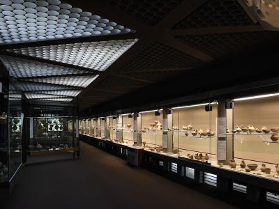 A long museum gallery with glass display cases filled with ancient pottery, illuminated by distinctive ceiling panels made of numerous small circular lights.