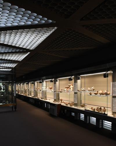 A long museum gallery with glass display cases filled with ancient pottery, illuminated by distinctive ceiling panels made of numerous small circular lights.