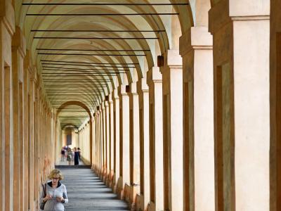 A long, covered arcade with repeating arches and columns, featuring a woman walking in the foreground and other pedestrians further down the path, under a vaulted ceiling with horizontal black cables or wires.