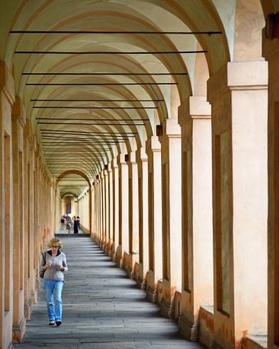 A long, covered arcade with repeating arches and columns, featuring a woman walking in the foreground and other pedestrians further down the path, under a vaulted ceiling with horizontal black cables or wires.