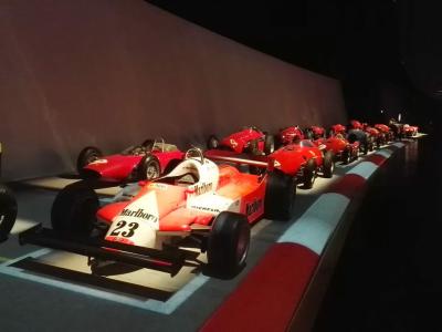 A row of red Formula 1 race cars (including a prominent Marlboro-liveried car with number 23) displayed on a track-themed floor inside a dimly lit museum gallery.
